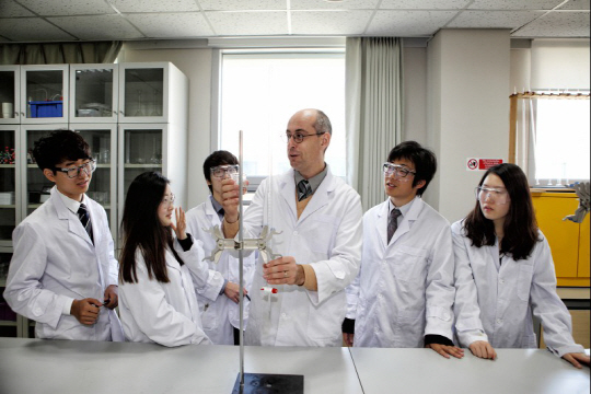 NLCS 학생들이 화학 시간에 실험 가운과 보안경을 쓰고 선생님과 함께 화학 실험을 하고 있다.