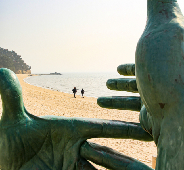 배미꾸미 조각공원에 설치된 손을 형상화한 대형 조각품 사이로 보이는 해변을 여행객들이 걷고 있다. 박주기부터 배미꾸미 조각공원까지 이어지는 구간은 모도에서도 가장 인기 있는 걷기 구간이다.