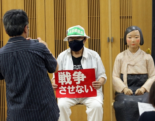 지난 24일 일본 교토시의 한 전시회에서 관람객이 ‘전쟁하게 하지 않는다’는 메시지가 적힌 종이를 들고 소녀상 옆에 앉아 기념사진을 찍고 있다. /연합뉴스